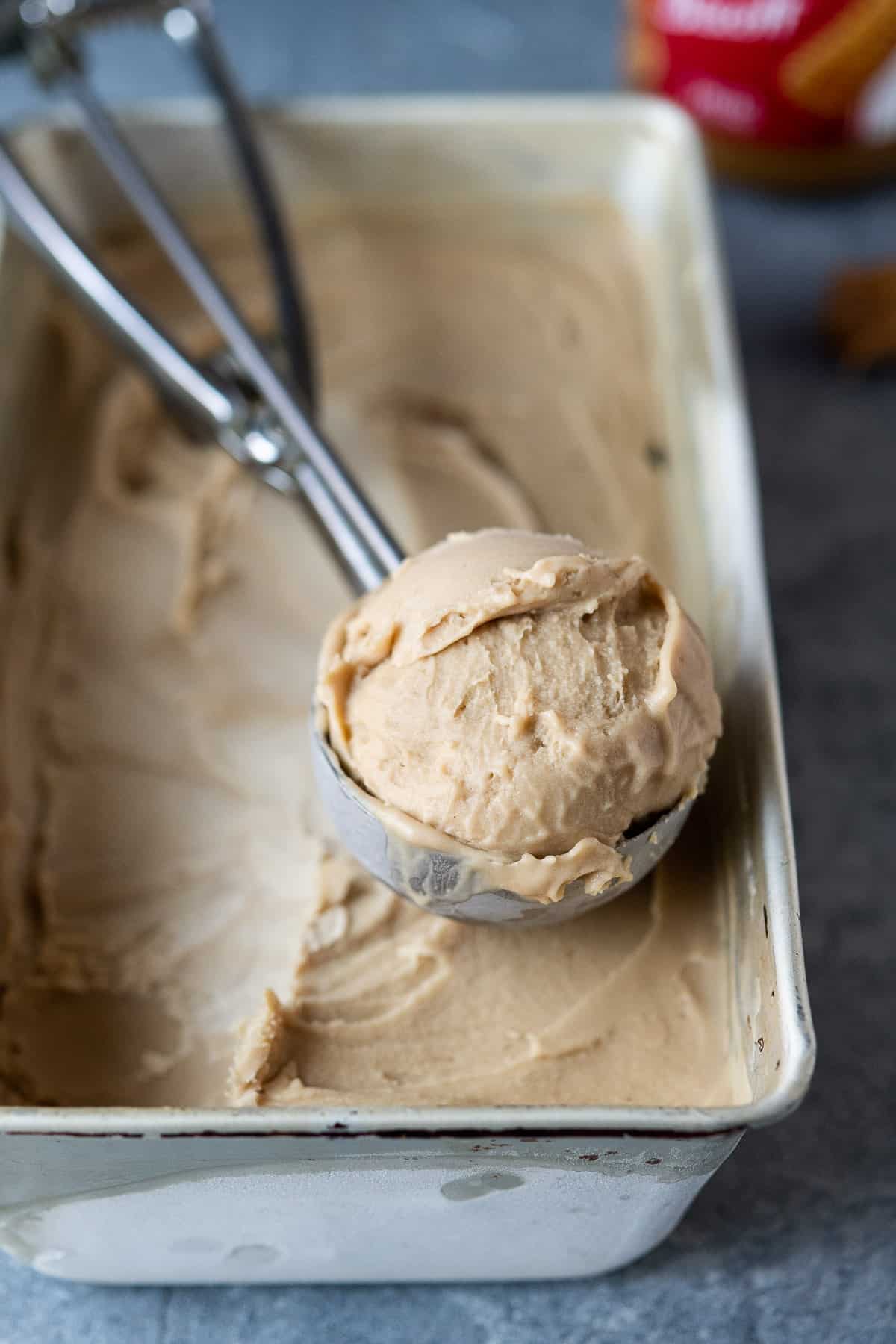 A metal loaf tin of vegan biscoff ice cream with an ice cream scoop and a jar of Biscoff.